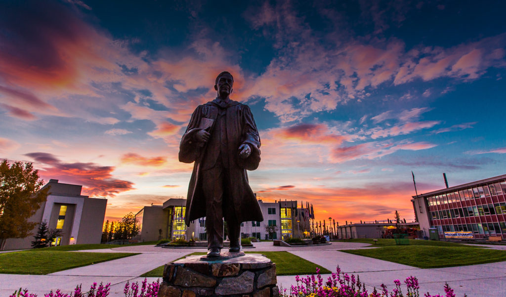 The Bunnell statue at the University of Alaska Fairbanks with a sunrise behind and flowers in front.