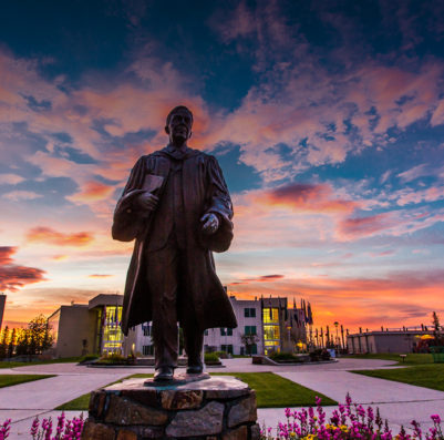 The Bunnell statue at the University of Alaska Fairbanks with a sunrise behind and flowers in front.
